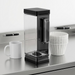 A Keurig K-2500 3.25L clear coffee brewer reservoir with a black lid is shown upright on a stainless steel counter next to a stack of classic paper coffee filters. A standard mug and pen are nearby, clearly demonstrating its considerable size and capacity for a busy environment. Clean product lighting on a white background.