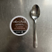 A top-down view of a Laura Secord Hot Chocolate K-Cup, exactly as seen in [image_0.png], placed neatly on a kitchen counter next to a common stainless steel teaspoon for scale. The lighting is soft and even.