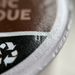Extreme close-up macro photograph of the Keurig Canada Inc. LAURA SECORD HOT CHOCOLATE-K CUP foil lid texture and printed details. The detailed white script, bilingual text, and recycling logo are sharp with minimal depth of field. The foil sheen is visible beneath. Neutral light.